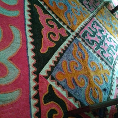 The image shows a close-up of a colorful felt rug with traditional Kyrgyz patterns. The rug features a vibrant pink border on the left, with a turquoise spiral design outlined in orange thread. The central part of the rug is divided into diamond-shaped sections. One section displays a black background with a pink stylized motif outlined in gold. Adjacent sections feature a light blue background with an orange, swirling pattern, also outlined in pink. Another section exhibits a dark gray background with pink and orange swirling designs. The overall impression is of intricate craftsmanship and cultural artistry. The rug is likely displayed indoors, possibly in a home in the village of Тосор, located in the Issyk-Kul Region of Kyrgyzstan, known for its traditional crafts and nomadic heritage. The lighting suggests an indoor setting, and the perspective indicates the rug is laid out flat, or possibly draped over a surface.