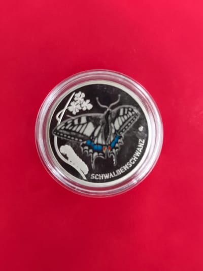 The image shows a close-up, overhead view of a round coin encased in clear plastic, resting on a bright red, uniform surface. The coin features a detailed engraving of a swallowtail butterfly, with intricate black and white wing patterns and hints of blue and red on its body. To the left of the butterfly, there is a stylized depiction of a caterpillar and some small flowers. Along the bottom edge of the coin, the text "SCHWALBENSCHWANZ" is visible, which translates to "swallowtail" in German. The coin appears to be a collectible item, possibly a commemorative piece related to local wildlife or a specific event. The red background is smooth and reflective, suggesting a polished tabletop or similar surface. There is no indication of the city of Triberg im Schwarzwald, Germany, in the image itself or any context provided.