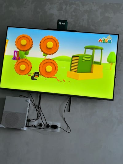 A television screen displays a cartoon scene of a green tractor on a bright green field under a blue sky. To the left of the tractor, there are three large orange cartoon wheels with yellow centers, each mounted on green cartoon stems. A purple and orange structure resembling a slide or playground equipment is partially visible behind the leftmost wheel. Above the tractor, a small digital clock displaying "09:48" is attached to the top edge of the television. To the right of the tractor, a simplified green tree is depicted. Below the television, a white game console is connected to a power strip with multiple electrical outlets, and various black cables are routed behind the console. The television is mounted on a textured gray wall. The scene appears to be an interior domestic setting in Dzerzhinsky, Russia.