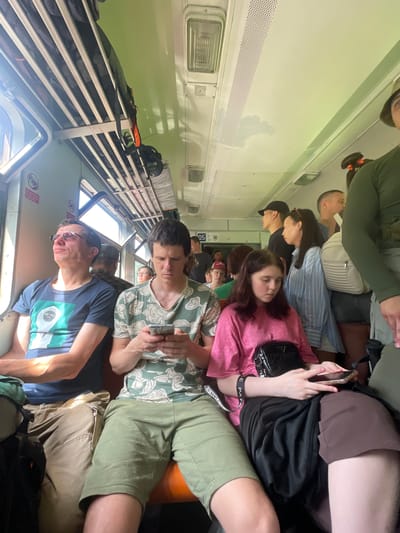 The image shows the interior of a train carriage, likely in Kuzmolovsky, Russia, with several passengers seated. The carriage has overhead luggage racks and visible windows. A man in a blue t-shirt and glasses is looking up and to the left. Next to him, a man in a green and white patterned t-shirt is looking down at his phone. A young woman in a pink shirt is seated in the foreground, also looking down at her phone, with a black purse on her lap. Other passengers are visible in the background, some standing and some seated, in various states of engagement. The lighting suggests it is daytime.