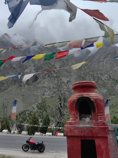 The image depicts a roadside scene in Jispa, India, characterized by prayer flags strung across the frame against a backdrop of rugged, cloud-shrouded mountains. A red motorcycle is parked on the shoulder of an asphalt road. In the foreground, a weathered, red, chorten-like structure stands prominently. The mountain slopes are sparsely covered with vegetation and rocks. The sky is overcast and hazy, contributing to the muted colors of the landscape. The prayer flags are of various colors, including blue, white, red, green, and yellow, and are fluttering in the wind. The red motorcycle is a modern design with a black seat and black wheels. The chorten structure has a hollow interior and appears to be made of concrete or brick, with visible signs of age and wear. The overall impression is one of a remote, spiritual location.
