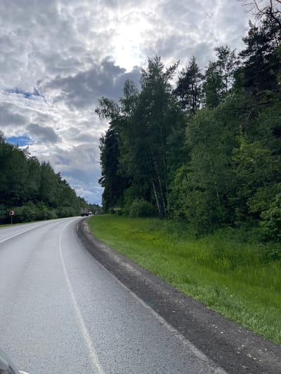 The image presents a rural road in Semyonovskoye, Russia. The asphalt is smooth, and the median stripe is painted white. The roadside has lush green grass and a thick forest line. The sky is overcast with heavy cloud cover, although the sun is still visible. There is little traffic on the road. The trees and the presence of clouds create an environment of overcast conditions.