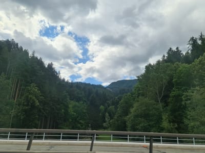 The image depicts a view from a bridge overlooking a lush, tree-covered valley with mountains in the background. The sky is partly cloudy with patches of blue visible. The bridge railing is in the foreground, with a road surface below. This scene is consistent with the landscape found near Leoben, Austria, characterized by its mountainous terrain and dense forests. There are no people or specific actions visible in the image. The environment is natural and serene.