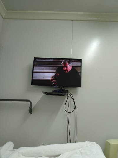 The image shows a television screen displaying a scene from a movie or TV show, with two men visible. The television is mounted on a light-colored wall. Below the television, a black shelf holds a digital receiver or set-top box, with several black cables dangling down. The wall appears to be paneled. To the left of the television, a metal railing is partially visible. In the foreground, a white duvet or blanket is spread out. A label reading "220 V" is affixed to the wall near the bottom of the frame. The environment appears to be an indoor space, possibly a bedroom or a simple living area. The location is specified as Қопа ауылдық округі, Kazakhstan.
