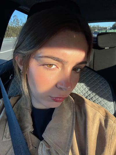 The image shows a woman in a car, possibly in Pombal, Portugal, although the exact location cannot be determined from the image. She is wearing a brown suede jacket and a dark-colored sweater or top underneath. A seatbelt is visible across her body. The interior of the car has quilted gray seats. There is bright sunlight casting shadows on her face. The woman has light skin, brown eyes, and glossy lips. The background shows a glimpse of trees and a blue sky through the car window.