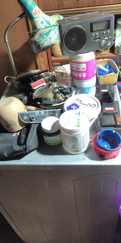 The image shows a tabletop or shelf in what appears to be a cluttered storage area, possibly a workshop or garage, in Есипово, Russia. On the surface, there are several items, including a small portable radio, various containers of what look like cosmetic or cleaning products, a black bag, a metal device with multiple inputs and outputs, and a red and blue electrical junction box. The background shows wooden shelves and a dimly lit environment. The overall impression is one of disarray and storage of miscellaneous items. There are no people or animals visible in the image.