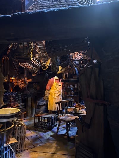 The image depicts the cluttered interior of what appears to be a workshop or storage area, possibly related to fishing or maritime activities, given the abundance of nets, ropes, and wooden crates. The scene is dimly lit, with strong shadows and a warm glow suggesting artificial lighting, possibly from lamps or the setting sun. The overall atmosphere is one of industriousness and perhaps a bygone era, with a rich texture of wood, rope, and woven materials.
The central subject is a figure dressed in a light-colored apron over a reddish garment, suggesting they are engaged in some form of manual labor or craft. This person is positioned amidst a chaotic yet organized collection of objects. Visible are various types of fishing nets, coiled ropes of different thicknesses, and numerous wooden crates and baskets stacked and piled throughout the space. Some of the crates appear to be crab or lobster pots. Shelves in the background are laden with pottery or jars, further adding to the sense of a working environment.
The setting is rustic and appears to be indoors, with wooden plank flooring and rough-hewn wooden beams and supports visible overhead. The textures of the materials, from the rough stone-like wall on the left to the weathered wood, contribute to a sense of authenticity and age. The lighting creates dramatic contrasts, highlighting certain areas and leaving others in deep shadow. The scene is likely a staged or historical representation, possibly within a museum or attraction, given the meticulous arrangement of the props and the theatrical lighting. No text is visible in the image. The location context of Watford, United Kingdom, does not provide specific clues within the image itself, but might suggest a heritage or historical site.