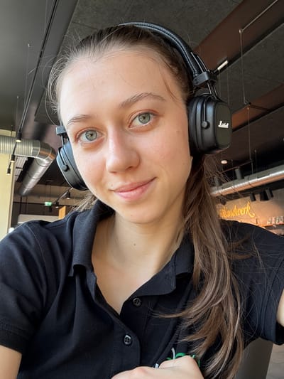This media file shows a close-up, rotated image of a young woman wearing black over-ear headphones. She has light skin, light brown hair, and light green eyes. She is wearing a black collared shirt. The background suggests an indoor setting, possibly a modern office or public space with industrial elements like exposed metal pipes and ceiling fixtures. The lighting appears to be artificial, indicating it could be daytime or evening.
The woman is looking directly at the camera with a neutral expression. The headphones on her head are black and have the "Marshall" logo in white on the earcups. Some of her hair falls over her shoulders and the top of her shirt.
Visible text in the background, on a lit sign, reads "Handwerk" which translates to "Craftsmanship" or "Trade" in German. This, combined with the location context of Elsenfeld, Germany, suggests the setting might be related to a craft-oriented business or exhibition space.