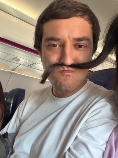 The image shows a close-up, selfie-style portrait of a man sitting on an airplane. He has brown hair and is wearing a white t-shirt. His mouth is pursed and he has a strand of dark hair across his upper lip, creating the appearance of a mustache. In the background, airplane seats and overhead compartments are visible. The scene is lit by artificial cabin lighting and natural light from a window. The man appears to be in motion, as indicated by the slight blur of his hair and the angle of the photograph. There is no indication of the city of Donja Cijevna, Montenegro in this image.