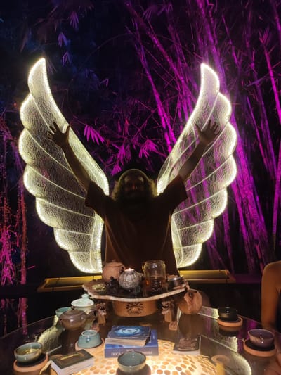 A man with long hair and a beard stands with his arms raised in front of illuminated wings. He is wearing a brown t-shirt. A table in front of him holds teacups, teapots, tarot cards, a candle, and other decorative items. The table is on a surface that is illuminated from below. Behind the man and wings is a backdrop of bamboo stalks lit with purple light. The scene suggests an outdoor setting at night, possibly in a decorative garden or tea ceremony area in Lokaserena, Indonesia.