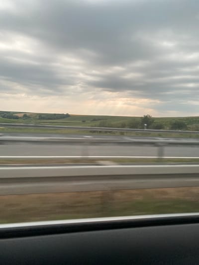 This media file captures a view from a vehicle, likely a train or bus, traveling through a rural landscape in Indjija Municipality, Serbia. The scene is oriented vertically, with the horizon running horizontally across the middle of the frame.
The dominant subject on the left side is a dramatic sky filled with heavy, grey clouds. Patches of sunlight break through the clouds, casting rays of light onto the landscape below. This suggests it might be late afternoon or early evening, with the sun low in the sky. The weather appears to be overcast with intermittent sunshine, creating a somewhat dramatic or moody atmosphere.
The right side of the frame is dominated by parallel lines, suggesting the interior of the vehicle, possibly window frames or a partition. Beyond these, the landscape unfolds. A road, possibly a highway or a railway line, is visible, running parallel to the vehicle's path. The land beside the road is a mix of green fields and trees, indicating a rural or semi-rural setting. A lone tree stands in the distance on a grassy slope, with a small white signpost nearby. The overall impression is one of travel through a scenic, perhaps agricultural, region. There is no visible text in the image.