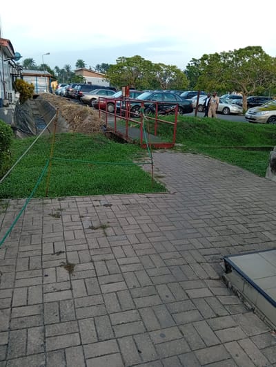 The image depicts an outdoor scene in what appears to be a parking lot or public area in Omagwa, Nigeria. The foreground is a paved walkway made of rectangular blocks. To the left, a narrow irrigation ditch separates the walkway from a building. A small, red metal bridge spans the ditch. Green grass covers the banks of the ditch, and a rope barrier runs along the edge of the walkway. In the background, several parked cars are visible, and two men in light-colored uniforms or suits are walking along the edge of the parking lot. Trees are scattered throughout the scene, providing some shade. The sky is overcast.