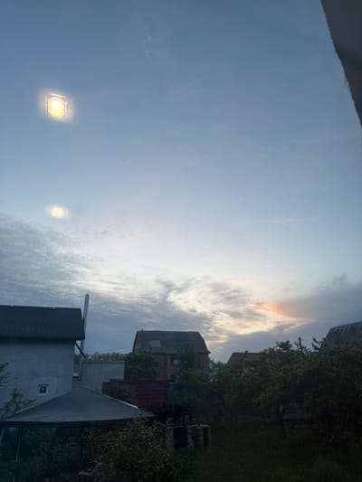 The image shows a view looking up towards the sky in Machukhy, Ukraine. The sky is predominantly blue with scattered clouds, some tinted with orange hues. There are buildings and trees visible on the right side of the frame. Two bright, square-shaped lights are visible in the sky.