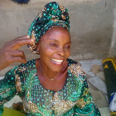 An adult female, likely middle-aged or older, with a dark complexion, is depicted in a medium shot within an indoor or semi-outdoor setting in Ibreda, Nigeria. She is smiling broadly, revealing white teeth, and her head is slightly tilted. She wears a patterned headwrap and a matching long-sleeved garment in green, black, and gold hues with geometric and abstract designs. A thin gold-colored necklace with a small pendant is visible around her neck. A dark-skinned hand, showing two extended fingers, is positioned near the left side of her head. The background consists of a plain, light grey concrete wall. To the woman's right, a tiled floor is visible, upon which rests a small, light brown cylindrical container with a lid, and a partially rolled or folded dark green and yellow patterned mat. Illumination is consistent with bright, natural light.