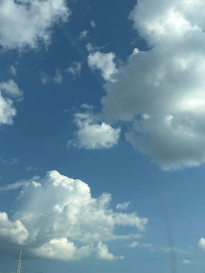 The image depicts a bright, daytime sky filled with white, fluffy cumulus clouds against a clear blue backdrop. There are no people or specific urban landmarks visible, with the exception of a portion of a metal electricity pylon and some power lines in the lower left corner, suggesting an outdoor, possibly rural or suburban environment. The clouds are varied in size and density, with some catching the sunlight and appearing luminous, while others are more shaded. The lighting indicates a sunny day, likely mid-morning or afternoon. The scene is static, with no discernible action or interaction. The location in Verinsko, Bulgaria, cannot be confirmed or denied from the visual information alone.
