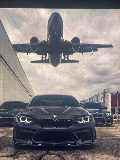 An airplane is captured in mid-flight, descending towards a runway. Below the aircraft, three dark-colored BMW cars are parked on an asphalt surface. The car in the foreground is a dark gray BMW M2, prominently displaying its distinctive headlights and front grille. To the left and right of the M2, two other BMWs are visible, partially obscured. The environment appears to be an industrial or airport setting, with a large building to the left and a chain-link fence in the background. The sky is overcast, suggesting a cloudy day. The scene does not contain any people or specific information about Paczkowo, Poland.