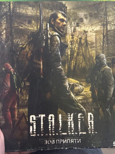 The image depicts the cover of the video game "S.T.A.L.K.E.R.: Call of Pripyat." The cover features two armed men amidst a desolate, overgrown urban landscape. One man lies prone, holding a sniper rifle. Another figure, wearing a gas mask, is seen further away, standing near a damaged building. The vegetation is thick and the buildings appear dilapidated. The background suggests a post-apocalyptic setting. The location is not in the city of Chuprino, Russia, but Pripyat, Ukraine.