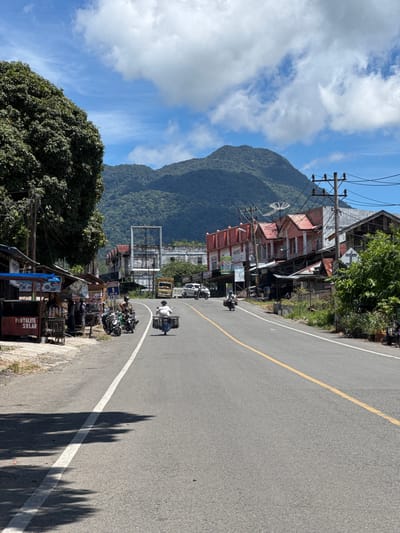 This is a medium-angle, eye-level shot of a road in Saree, Aceh, Indonesia, on a bright, sunny day with scattered clouds. The road curves slightly to the right and slopes gently uphill. On the left side of the road, there are several small shops and stalls, some with blue awnings, and a large tree with dense foliage. A sign on one of the stalls reads "PERTALITE SOLAR". Several motorcycles are parked near these shops.

Further down the road, on the right, are more buildings, some with red-tiled roofs and others appearing more modern. There are utility poles with numerous wires running overhead. In the background, lush green mountains rise against the blue sky.

There are several people and vehicles visible. A person on a motorcycle with a sidecar is traveling away from the camera in the right lane. Two people on motorcycles are stopped or moving slowly near the shops on the left. Another person is seen walking on the sidewalk on the left. In the distance, a yellow truck and a white car are on the road. The overall scene depicts a lively street in a mountainous region.