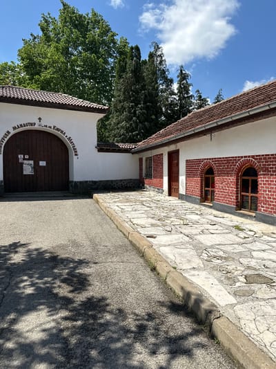 The image shows the exterior of the Maleevski Monastery "St. St. Cyril and Methodius" in Barziya, Bulgaria. The monastery building features a red brick and white facade, with arched windows and a wooden door leading into the structure. A stone pathway runs alongside the building, bordered by an asphalt road with tree shadows cast upon it. The monastery entrance is marked with inscription above the door. Trees and blue sky with clouds are visible in the background.
