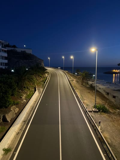 The image presents an elevated view of a winding road illuminated by streetlights at night. The road appears freshly paved, marked with solid white lines on either side and a central dashed white line.
On the right side of the road, a metallic guardrail is present along with natural terrain including grass and some small bushes. Palm trees are visible in the distance.
On the left side, the land slopes downwards, revealing a view of buildings, a rock face, and more greenery. In the distance, across the water, buildings with lights are visible, suggesting a coastal settlement.
Based on visual details, specifically the architecture and coastal setting, the location can be identified as Puerto Rico, Gran Canaria, Spain.