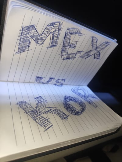 The image shows an open notebook with blue ink drawings of letters on lined paper. The top page has the letters "MGK" drawn in large, blocky, shaded capital letters. Below this, on the same page, the letters "US" are visible, with the "U" and "S" appearing smaller and partially obscured. The bottom page displays the letters "YO", with the "Y" drawn in a large, blocky, shaded capital letter and the "O" drawn as a more abstract, rounded shape with shading. The notebook is angled, with the top page bent slightly upwards, revealing the bottom page. The background is dark. The image does not depict any specific location in Thôn 18, Vietnam.