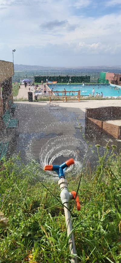 In Сырымбет, Kazakhstan, an outdoor scene unfolds under a partially cloudy sky. In the foreground, a garden hose nozzle, a blue Y-shaped sprinkler with orange tips, is actively spraying water in a circular pattern onto a paved area. The spray creates a mist that catches the sunlight. Lush green grass and some flowering plants surround the nozzle.
Beyond the immediate foreground, a paved walkway leads towards a swimming pool area. Several people are visible, some relaxing by the pool and others in the water. There are lounge chairs and umbrellas set up. A dark trash receptacle is situated near the walkway. To the left, a building with a textured, light brown facade is partially visible. In the background, a fence encloses the pool area, and beyond that, rolling hills covered in green vegetation extend towards the horizon. The overall impression is a recreational area on a sunny day, with the sprinkler suggesting a method of cooling or watering the surroundings.