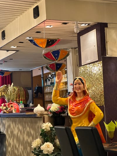 The interior of a restaurant in Saint-Josse-ten-Noode, Belgium, located at 50.848 latitude, 4.374 longitude. The scene features a life-sized mannequin of a woman wearing a traditional Indian outfit. The woman is standing on a countertop with her right hand raised to her forehead, as if she is waving. The background is adorned with colorful umbrellas hanging from the ceiling. The scene is lit by overhead lights, and there are various objects on the countertop, including a plant and a white ceramic object. The restaurant appears to be decorated in a traditional Indian style. There are also a few chairs placed in front of the mannequin, and the countertop appears to be made of wood. The walls are painted in warm, earthy tones. The image suggests that the restaurant is a popular spot for people seeking authentic Indian cuisine. The overall tone of the image is cheerful and welcoming.