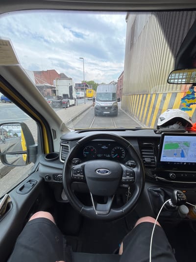 This is a first-person perspective, point-of-view shot taken from inside the driver's cabin of a yellow Ford Transit van, driving through the city of Neuss, Germany. The driver's legs, clad in black shorts, are visible in the foreground. The steering wheel, dashboard, and a large touchscreen infotainment system displaying a navigation map are prominent. Outside the van, a narrow street or industrial area is visible, with buildings on either side. A white semi-trailer truck is parked or moving ahead of the van. The weather appears to be overcast with a cloudy sky. The navigation map on the screen indicates the current location as Neuss, with "Rennbahn Park" and "Hammer Landstraße" labeled on the map.