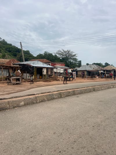 The image depicts a street scene in Minda, Nigeria. On the left side of the frame, a row of small, simple structures with corrugated metal roofs lines the side of a paved road. These appear to be shops or stalls, some with open fronts displaying goods or wares. A few people are visible standing or walking near these buildings, engaged in everyday activities.
In the foreground, a man in a grey t-shirt and shorts stands near a wooden structure, appearing to be interacting with someone or something out of frame. Further down the street, several other individuals are scattered amongst the stalls. The setting suggests a typical day in a community, possibly in the morning or afternoon given the lighting. The sky is overcast with grey clouds.
The road itself is paved but shows signs of wear, with a concrete curb bordering the shops. Beyond the buildings, lush green trees and a hillside are visible, indicating a somewhat rural or semi-urban environment. Power lines stretch across the sky above the buildings and trees. There is no visible text in the image. The overall impression is one of a bustling, yet modest, community street.