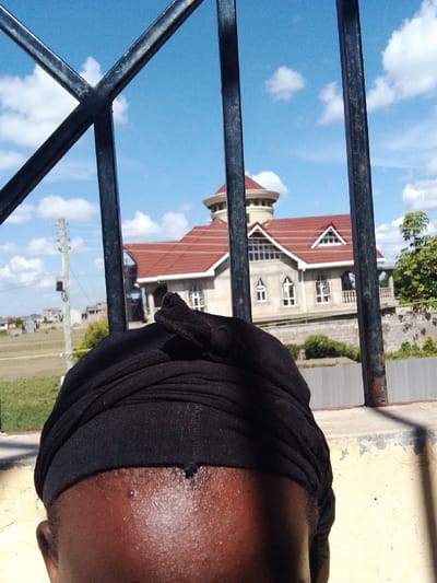 The image depicts an outdoor scene in Kalimoni ward, Kenya, viewed from behind a black metal barred structure and a yellowish concrete ledge. In the immediate foreground, the top of a dark-skinned individual's head, partially covered by a black headwrap, is visible. The forehead shows a slight sheen. Through the metal bars, a large, multi-story building with a prominent red tiled roof is centered in the midground. This building features multiple gables, white-framed windows, and a distinctive light-colored cylindrical turret structure on its upper left side. Below the building, a grey retaining wall is present. To the left of the building, an open grassy field with scattered smaller structures and a utility pole is visible. Lush green trees are seen on the right side of the building and in the distant background. The sky is bright blue with scattered white cumulus clouds, indicating a clear, sunny day. No explicit actions or interactions are visible.