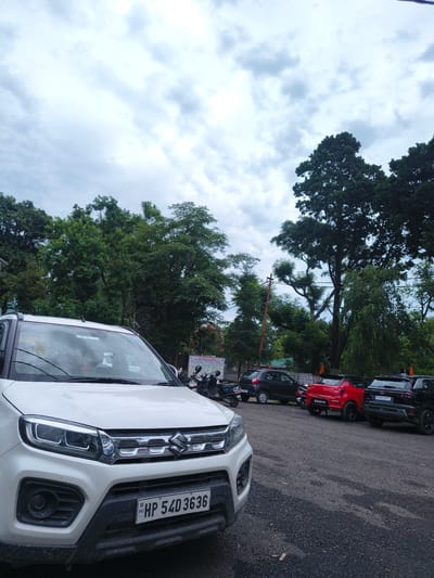 The image shows a parking lot in Chelian, India, under a cloudy sky. In the foreground, a white Suzuki Brezza SUV is parked, facing towards the viewer. The vehicle's license plate reads "HP 54D 3636". Behind the SUV, several other vehicles are visible, including a red Maruti Suzuki Swift and a black Ford EcoSport. A row of motorcycles is parked to the left of the red Swift. The parking lot is paved with asphalt. Lush green trees form a dense backdrop, with some taller trees partially obscuring the sky. A sign with text is visible to the right of the motorcycles.