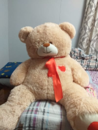 A close-up, angled shot shows a large, plush teddy bear lying on a surface, possibly a bed or sofa. The bear is light brown with a white snout, dark button eyes, and a small orange nose. It wears a bright red ribbon tied around its neck, with a red heart appliqué visible on its chest just above the ribbon. The background is blurred, with hints of patterned fabric, including plaid designs in shades of gray, white, and purple, suggesting a domestic setting. The lighting is soft, and the overall mood is relaxed and cozy. No text is visible in the image. The location context provided is Khodorkivtsi, Ukraine.