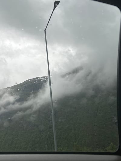 This media file depicts a view from inside a vehicle, likely a bus or train, looking out a window. The main subjects are a mountainous landscape partially obscured by clouds and fog, and a visible portion of the vehicle's interior, including a section of its frame and a lamppost extending from the left side of the frame.

The scene is set in a mountainous region, with steep, green slopes covered in vegetation. Clouds and mist are actively swirling around the mountains, creating a dramatic and somewhat moody atmosphere. Some patches of snow are visible on the higher elevations of the mountains, suggesting a cooler climate or season. A thin stream or road appears to wind down the mountainside in the distance.

The weather appears overcast and potentially damp, with small droplets or smudges visible on the window, indicating rain or mist. The lighting is diffused and subdued, consistent with a cloudy day. The time of day is indeterminate due to the heavy cloud cover.

The lamppost, a prominent object in the foreground, is metallic and extends diagonally across the frame, with a black light fixture at its end. It appears to be a street lamp or a similar external light source. The vehicle's interior is only hinted at by the edge of the window frame on the right side of the image.

There is no visible text in the image. The overall impression is one of natural beauty encountered during travel, with the weather adding a sense of mystery and grandeur to the mountainous environment of Røldal, Norway.