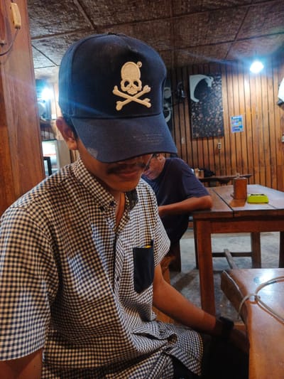 The image depicts two men seated in a rustic, wood-paneled indoor setting, likely a casual eatery or cafe in Geulumpangmeujim-Jim, Indonesia. The foreground features a man, seen from the chest up, whose face is mostly obscured by the brim of a dark blue baseball cap. The cap prominently displays a beige skull and crossbones emblem, with a small registered trademark symbol (®) above the skull. A mustache is visible beneath the cap's shadow. He wears a short-sleeved, black and white checkered shirt with a dark blue pocket. His right arm rests on a wooden table, where a white cable or wire is visible.
In the mid-ground, another man is partially visible, seated at a separate wooden table, wearing a dark t-shirt and glasses, and appears to be looking down. The setting is characterized by vertical wooden paneling on the walls and a textured, possibly woven, ceiling. Decorations include a crescent moon-shaped object and another dark, abstract artwork on the wall. A bright overhead light fixture is visible in the background, casting a noticeable glare and suggesting an evening or dimly lit interior. On the table in the mid-ground, a small green ashtray and a wooden salt or pepper shaker are present. The scene conveys a relaxed and informal atmosphere. Beyond the registered trademark symbol on the cap, no other explicit text is legible.