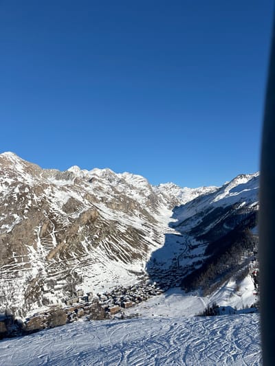 This vibrant, high-angle outdoor shot captures a majestic snow-covered mountain landscape under a brilliant, clear blue sky. The image appears to be taken from a ski lift, gondola, or elevated viewpoint, with a dark horizontal bar visible at the top (which would be a vertical bar on the left if rotated correctly to landscape orientation), suggesting part of the viewing structure.
The scene showcases the rugged beauty of the French Alps, specifically Val-d'Isère. Dominating the frame are towering, snow-dusted mountains, with exposed rocky faces revealing intricate geological patterns. Patches of dark evergreen trees cling to the steeper slopes, contrasting with the white snow and grey rock.
Towards the right side of the image, a wide, meticulously groomed ski piste is visible, crisscrossed with numerous ski tracks, indicating active skiing. Nestled further down, what appears to be a village or cluster of buildings, likely traditional chalets or resort facilities, is visible, their roofs blanketed in snow. Other ski infrastructure, such as potential lift towers, can be faintly discerned in the distance.
The bright sunlight and clear blue sky suggest a perfect winter day, ideal for outdoor activities like skiing. The overall atmosphere is one of serene grandeur and the excitement of a popular ski destination. No discernible text is visible in the image. People are implied by the ski tracks and resort setting but are not individually visible.
