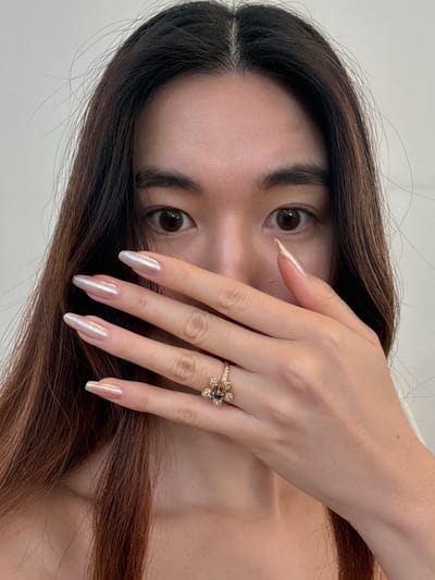 A close-up, head-and-shoulders portrait of a person, likely female, with long, dark brown hair parted centrally. The individual's dark brown eyes are open and directed towards the viewer. The left hand is raised, partially covering the lower half of the face, specifically the nose and mouth area. The fingers are extended, displaying long, almond-shaped nails with an iridescent, pearlescent light finish. A gold-toned ring, featuring a central dark blue or black gem surrounded by smaller clear stones arranged in a floral pattern, is worn on the fourth finger of the left hand. Exposed skin on the upper chest and shoulders indicates a garment with an open or wide neckline. The background is a uniform, plain light color, suggesting an indoor setting. This scene is located in Arcueil, France.