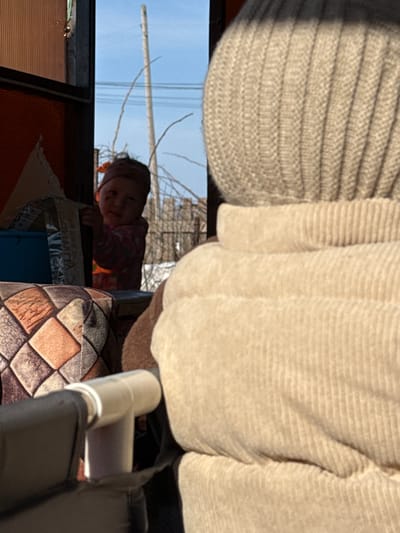 A low-angle shot from within a structure looks out to the sky and a young child. The child peers in, framed by a window or opening. Outside, bare tree branches are visible against a blue sky. A utility pole stands in the distance. Inside, a person wearing a light-colored, ribbed sweater or cardigan is positioned above the viewer, partially obscuring the view. A patterned cushion and a portion of a black and white structure with a plastic pipe are also in the foreground. The location is Кипчак, Russia.