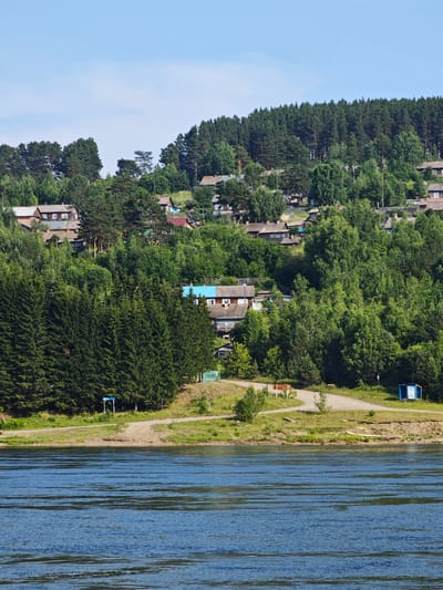 The image shows a river in the foreground, with a village on a forested hillside in the background. The village consists of wooden houses with various colored roofs, some of which are visible through the trees. The houses are situated on a steep incline, with a dirt road winding through the settlement. The river is calm and reflects the blue sky. The trees are dense and green, indicating a summer season. On the riverbank, there are some utility structures and signs, suggesting a populated area. The overall impression is of a rural community nestled in a natural landscape. This scene is likely located in or near the city of Pristan', Russia, given the typical architecture and natural environment of the region.