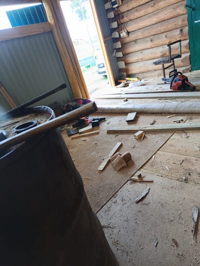 The image depicts the interior of a partially constructed wooden building, likely in Islambaevo, Russia. The walls are made of stacked logs, and the floor is covered with wooden planks and sawdust. In the foreground, a large, dark cylindrical object, possibly a barrel, is partially visible, with a metal rod resting on its rim. To the right, a red and black chainsaw sits on the floor next to a small, black metal stool. Various pieces of lumber and building materials are scattered around the floor. The entrance to the building is open, revealing a glimpse of the outdoors with a blue fence and trees. The scene suggests ongoing construction or renovation work. There are no people visible in the image.
