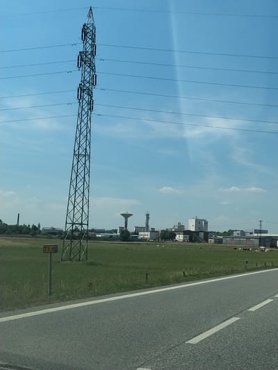 The image shows a rural landscape in Horažďovice, Czechia. In the foreground, there is a paved road with white lines. To the left of the road, a green metal electricity pylon stands tall, with several power lines extending horizontally across the frame. A yellow sign with "71.5" is mounted on a post next to the pylon. Beyond the pylon, a grassy field stretches out, dotted with some trees and various industrial buildings in the distance. One notable structure is a tall water tower. In the field, a few cows can be seen grazing. The sky is blue with scattered white clouds. The overall scene depicts an industrial area adjacent to a rural setting.