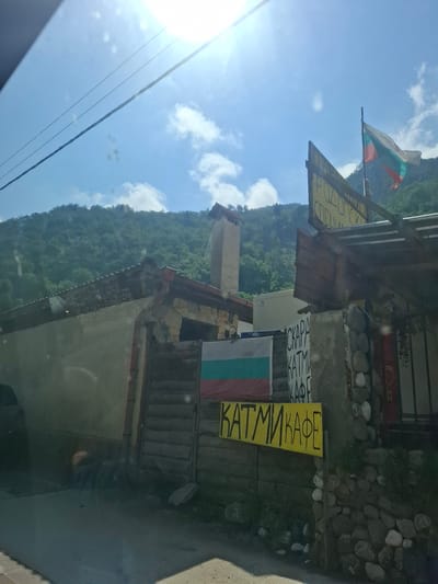 The image, taken on a bright, sunny day, depicts a mountainous landscape with a small commercial establishment in the foreground, likely in Нареченски бани, Bulgaria. The background features lush, densely forested green mountains under a clear blue sky with some scattered white clouds. The sun shines intensely from the upper left, creating a prominent lens flare.
In the mid-ground, a building with stone and concrete elements is visible on the right side of the frame. Two Bulgarian flags are displayed: a larger one waving from a pole against the sky, and a smaller one horizontally attached to the building's facade. Power lines cross the upper left quadrant of the image.
Several signs with Cyrillic text indicate the nature of the establishment. A partially visible yellow sign at the top reads "РОДОПСКИ СПЕЦИАЛИТЕТИ" (Rhodopean Specialties). Below it, a white sign displays "СКАРА" (Grill) and "КАТМИ" (Katmi). A larger, vertically oriented yellow sign prominently features "КАТМИ КАФЕ" (Katmi Cafe), suggesting the place is a cafe or restaurant specializing in grilled items and "katmi" (a traditional Bulgarian pastry/pancake).
No people are visible in the scene. The time of day is clearly daylight, likely midday, given the brightness of the sun. The setting appears rural or semi-rural, characteristic of a mountain village or resort area.