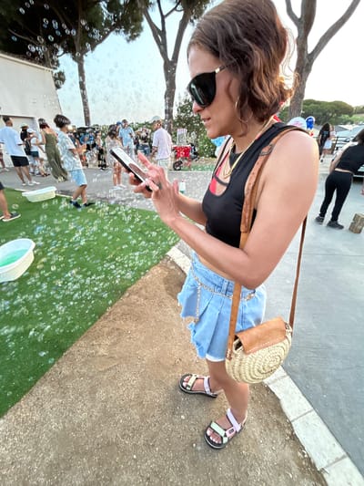An outdoor scene with many bubbles floating in the air. In the foreground, a woman is looking at her smartphone. She is wearing a black crop top, a denim skirt, and colorful sandals. She has a straw bag slung over her shoulder. In the background, other people are visible, some walking and some interacting with a bubble machine. There are trees and a building in the background, and the ground is a mix of gravel and artificial turf. This event is taking place in Sant Vicenç De Montalt, Spain.