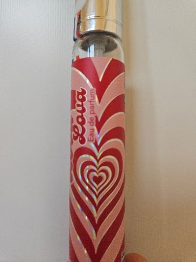 The image displays a clear glass cylindrical bottle containing "Lova Eau de parfum" against a plain, light-colored, vertically textured background. The bottle features a metallic silver-colored spray mechanism at its top. A label encompasses the majority of the bottle's body, characterized by a repeating pattern of concentric hearts. This pattern consists of alternating bands of dark pink/red and light pink/white, with some bands exhibiting a holographic or iridescent effect. The brand name "Lova" is rendered in a stylized, red, bubble-letter font and positioned vertically on a light pink section near the upper left of the visible label, followed by the vertically written text "Eau de parfum." A small, blurry, reddish-brown object is partially visible at the bottom right corner of the frame. No individuals, direct actions, or interactions are depicted. The immediate environment is sterile and devoid of specific identifying markers, preventing the determination of a precise location within Villeneuve-Saint-Georges, France, or any other specific geographic area from the visual evidence presented.