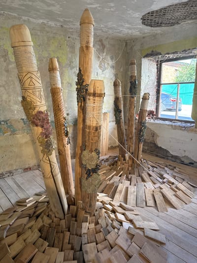 The image depicts an art installation composed of several large, carved wooden pillars of varying heights, placed in the interior of a dilapidated building. The pillars are adorned with intricate carvings, twine wrapping, and decorative metal elements resembling flowers and foliage. The floor is covered with scattered wooden planks, creating a dynamic base for the installation. The walls of the building are distressed, with peeling paint and exposed brickwork, and a damaged ceiling reveals the underlying structure. A window on the right side of the frame looks out onto an outdoor scene with a car and greenery. This artwork is located in the abandoned "Elec Electro-mechanical Plant" in Yelets, Russia, which has been repurposed as a space for contemporary art exhibitions.