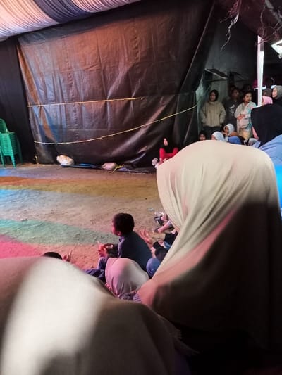 This image captures a gathering of people within a temporary tent-like structure in Kecamatan Cimaung, Indonesia. The walls of the enclosure are constructed from large, dark black tarpaulins, with a yellow rope stretched across one section.

In the foreground, the back of a person wearing a light brown or beige hijab dominates the right side of the frame. To their left, several children and adults are seated on the ground, facing forward towards an unpictured focal point. One child in a dark shirt is visible from behind, appearing attentive. The ground in this area is dirt, illuminated by patches of colored light, including red, blue, and green, suggesting the presence of stage lighting or a projector.

Further back, near the tarp wall, a group of individuals, mostly women and young people, stand and observe the event. Many are wearing head coverings, and one woman is seen in a light brown hooded jacket. On the far left, a stack of green plastic chairs is visible, and another woman in a red top and black hijab is seated on the ground near the tarp wall.

The scene depicts a community event or gathering, with participants actively watching or listening to something occurring outside the frame to the right. The temporary nature of the structure suggests a makeshift venue, suitable for various community activities, possibly in the evening or a heavily shaded daytime environment. No visible text is present.