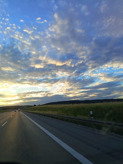 A low-angle, ground-level shot from a moving vehicle shows a highway in Dornstadt, Germany, under a vast, partially cloudy sky. The road is asphalt with clear white lane markings. A metal guardrail runs along the right side of the highway. Beyond the guardrail, a field of tall, dry grass stretches towards a dark, tree-lined hill in the distance. The sky is a mixture of deep blue and white clouds, with the setting sun casting a warm glow on the horizon, illuminating some of the clouds with golden and pink hues. Two trucks are visible on the highway in the distance.