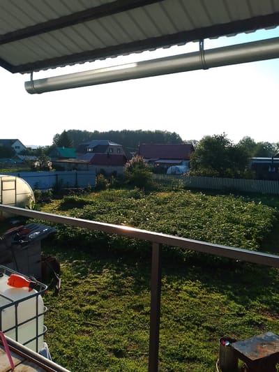 The image depicts a view from an elevated vantage point, likely a porch or balcony, looking out over a rural backyard or dacha property in Kranokholmskiy, Russia. The foreground features a dark metal railing and a portion of a corrugated metal roof with a gutter system.
Beyond the railing, a lush green lawn is visible, dappled with sunlight. To the left, a large white IBC tote with a black metal frame is partially visible, with an orange spray bottle resting on top. Further back, a greenhouse with a white, translucent cover stands amidst overgrown green vegetation.
A white picket fence runs across the middle ground, separating the yard from a collection of houses. These houses are varied in style and color, with some featuring red tile roofs and others lighter colored facades. Trees and foliage are abundant throughout the scene, forming a dense line of green along the horizon. The sky is clear and bright blue, indicating a sunny day. The overall impression is one of a peaceful, domestic setting in a Russian countryside.