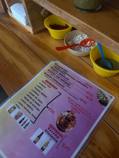 A tilted overhead shot shows a wooden table in what is likely a restaurant in Tepoztlán, Mexico. Arranged on the table are four yellow bowls, each filled with a different ingredient: one with a dark green powder, one with crumbled white cheese, one with dried red chilies, and one with green salsa. A blue spoon is in the bowl with the green powder, and an orange spoon is in the bowl with the cheese. A laminated menu lists different Mexican dishes and drinks with prices. The menu lists "Huaraches" for 135 pesos, "Sopes Sencillos" for 30 pesos, "Sopes con Guisados" for 35 pesos, "Sopes Especiales" for 45 pesos, "Rico Cafe de Olla" for 30 pesos, "Coca Cola" for 30 pesos, and "Cervezas Corona or Victoria" for 40 pesos. Next to the sodas section are images of various soda brands. Beside the bowls are a stack of folded white napkins. The angle and cropping focus on the food preparation elements rather than the broader environment.