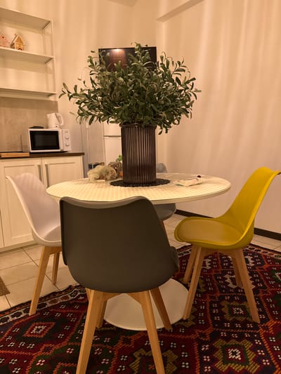 A still life photograph depicts an indoor scene with a dining area adjacent to a kitchen setting. The dominant feature is a round dining table surrounded by three chairs of varying colors (yellow, gray, and white), suggesting a modern design aesthetic. A large ornamental plant in a dark vase sits centrally on the table, partially obscuring a view of assorted packaged goods. In the background, there is a television and open shelving. A white microwave is visible on the countertop. The area is lit with warm light. A section of a patterned rug can be seen. There is no indication of location.