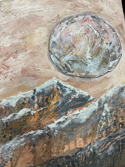 The image is a close-up, textured oil painting of a mountainous landscape under a large, textured orb, possibly a moon or sun. The sky is rendered in broad, streaky brushstrokes of pale pink and beige, with some areas of lighter white and subtle yellow. The orb itself is heavily textured, with thick impasto application of paint in shades of white, grey, pink, and gold, suggesting a rough, uneven surface. The mountains in the foreground are also depicted with heavy texture, featuring dark greys, browns, and oranges with streaks of white paint to represent snow or ice on the peaks. The overall impression is one of a dramatic and stylized natural scene. There are no people, animals, or specific structures visible, nor any indication of a city or location.